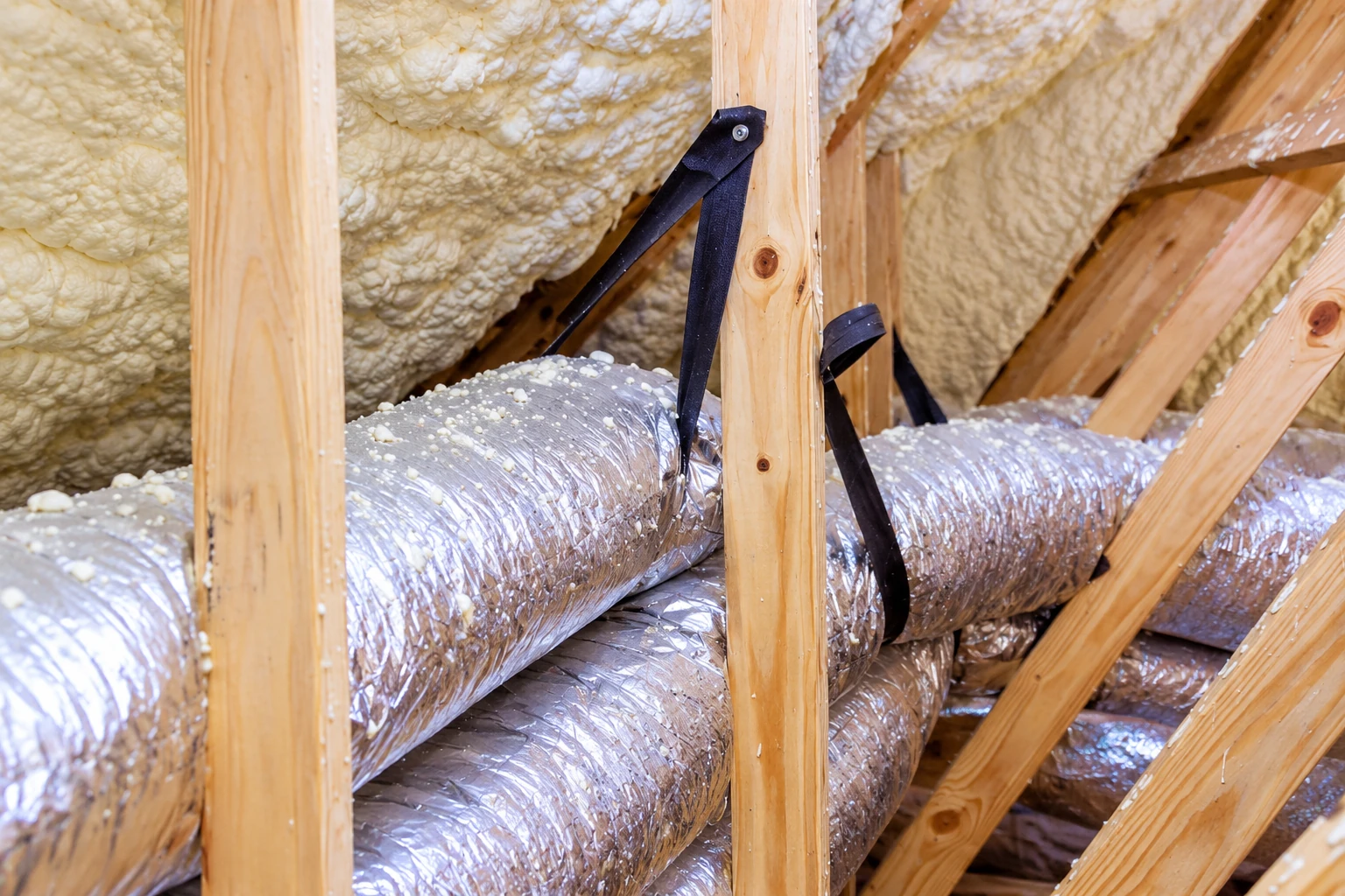 Foil-wrapped ducts run through an attic beneath spray foam insulation, secured among exposed wooden rafters.