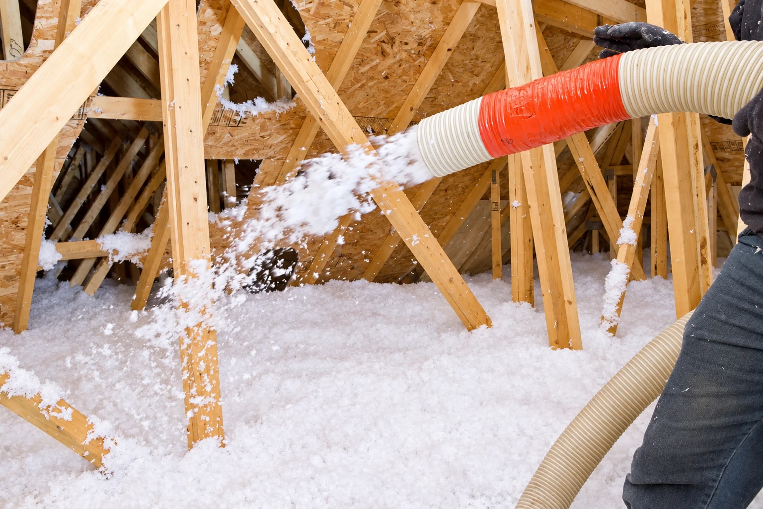 Blown-in insulation is sprayed from a flexible hose across the floor of a timber-framed attic.