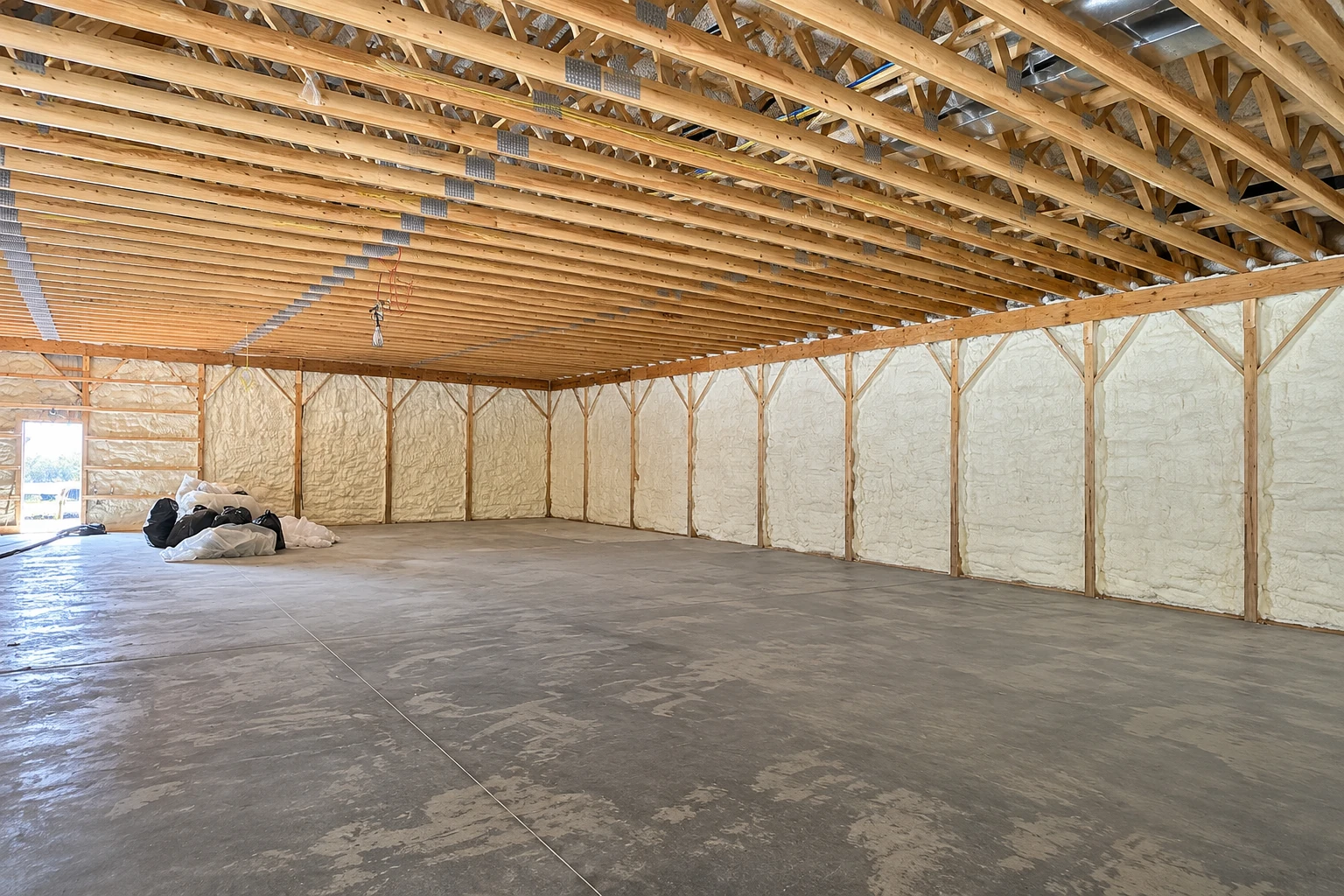 Large unfinished space with pale spray foam insulation covering framed walls beneath exposed wooden roof trusses.