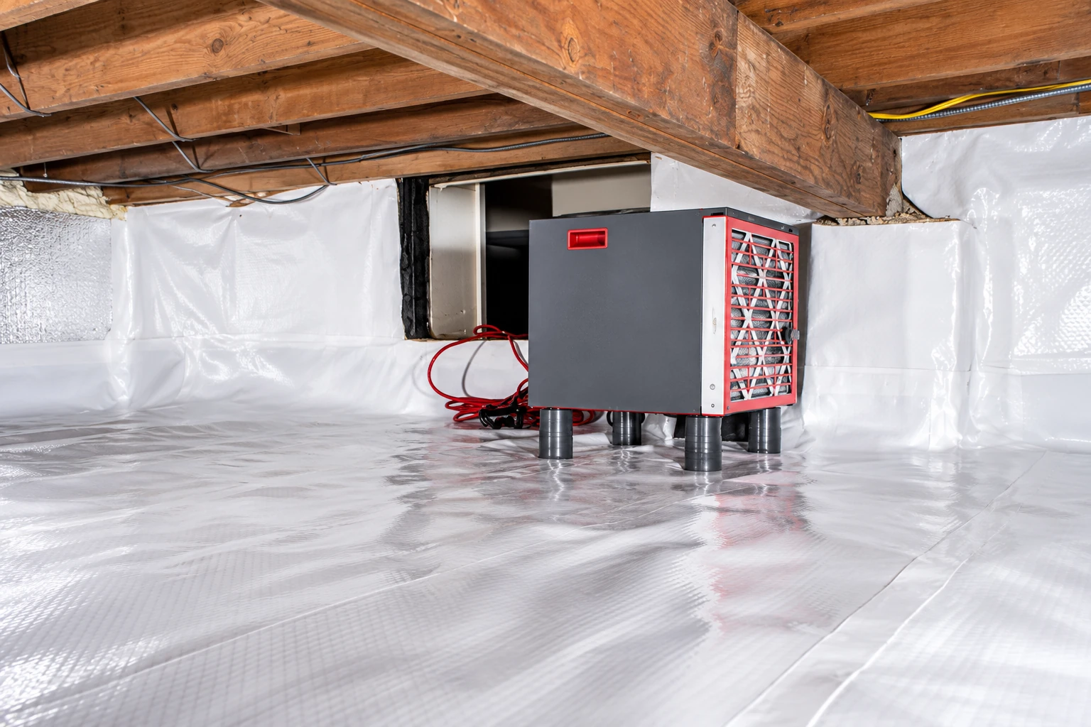 A gray dehumidifier stands on a plastic vapor barrier beneath exposed wooden beams in a crawl space.