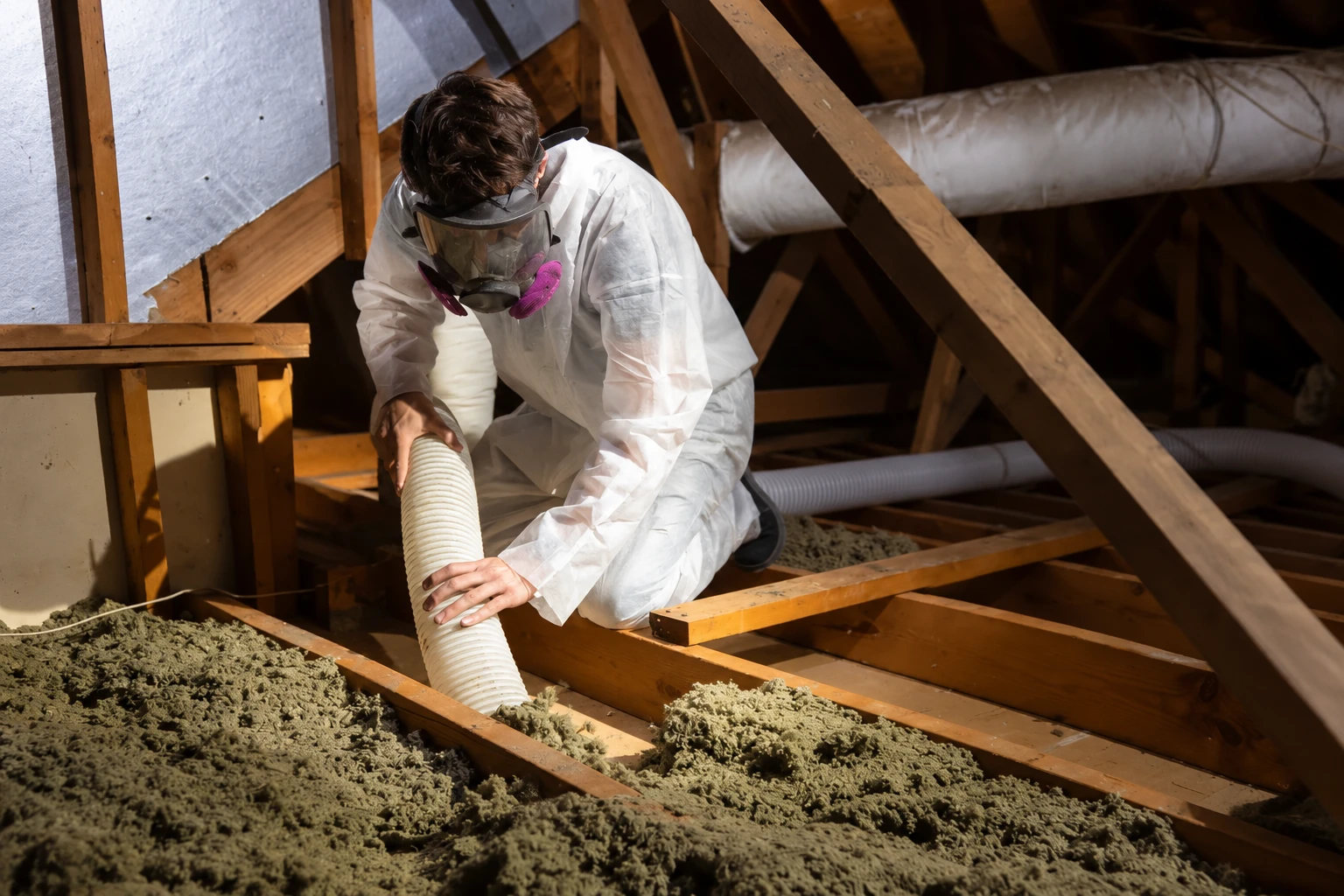 A worker in white protective clothing and a respirator uses a hose to remove loose attic insulation.